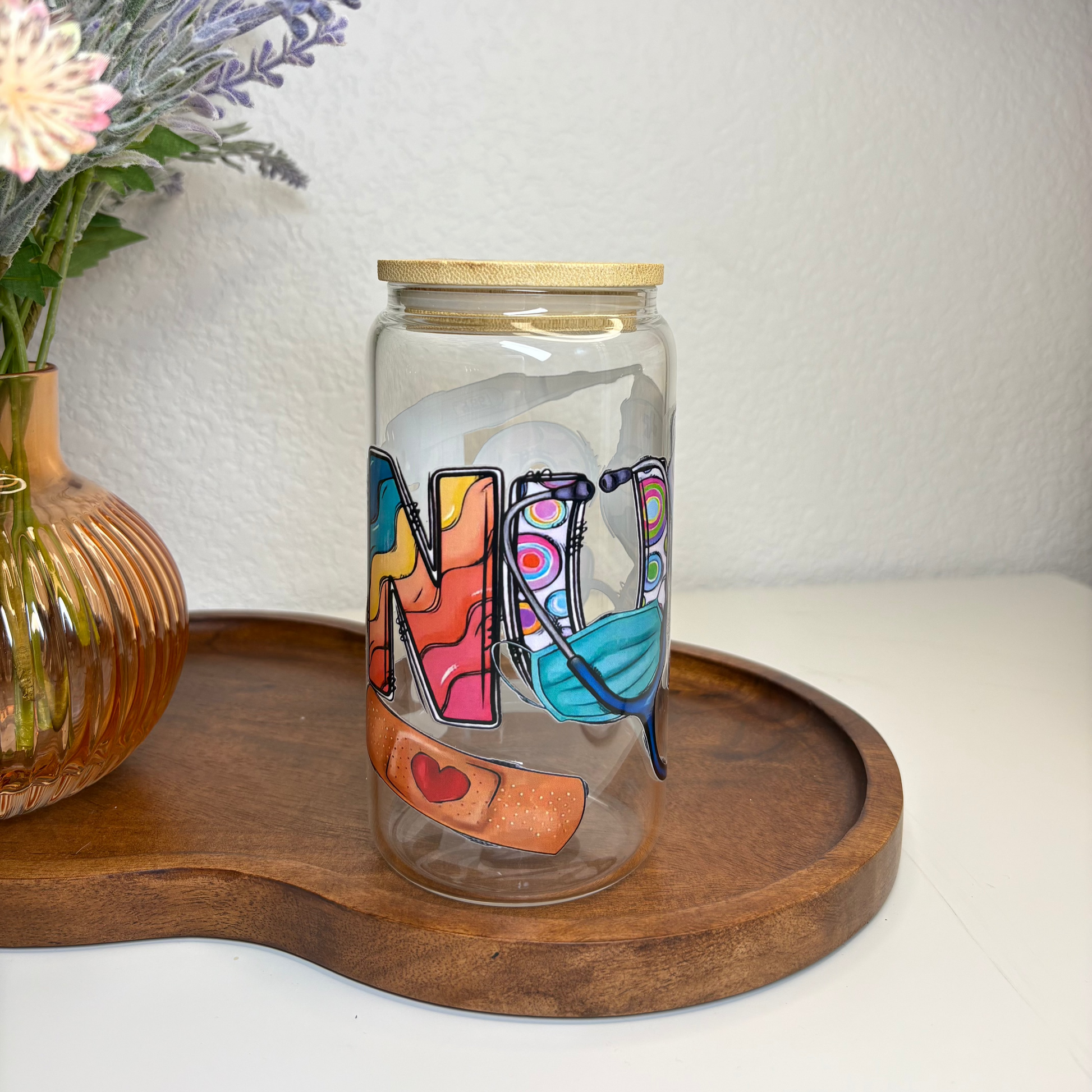 Decorative glass jar with colorful letters on a wooden tray, next to a vase with flowers.