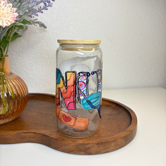 Decorative glass jar with colorful letters on a wooden tray, next to a vase with flowers.