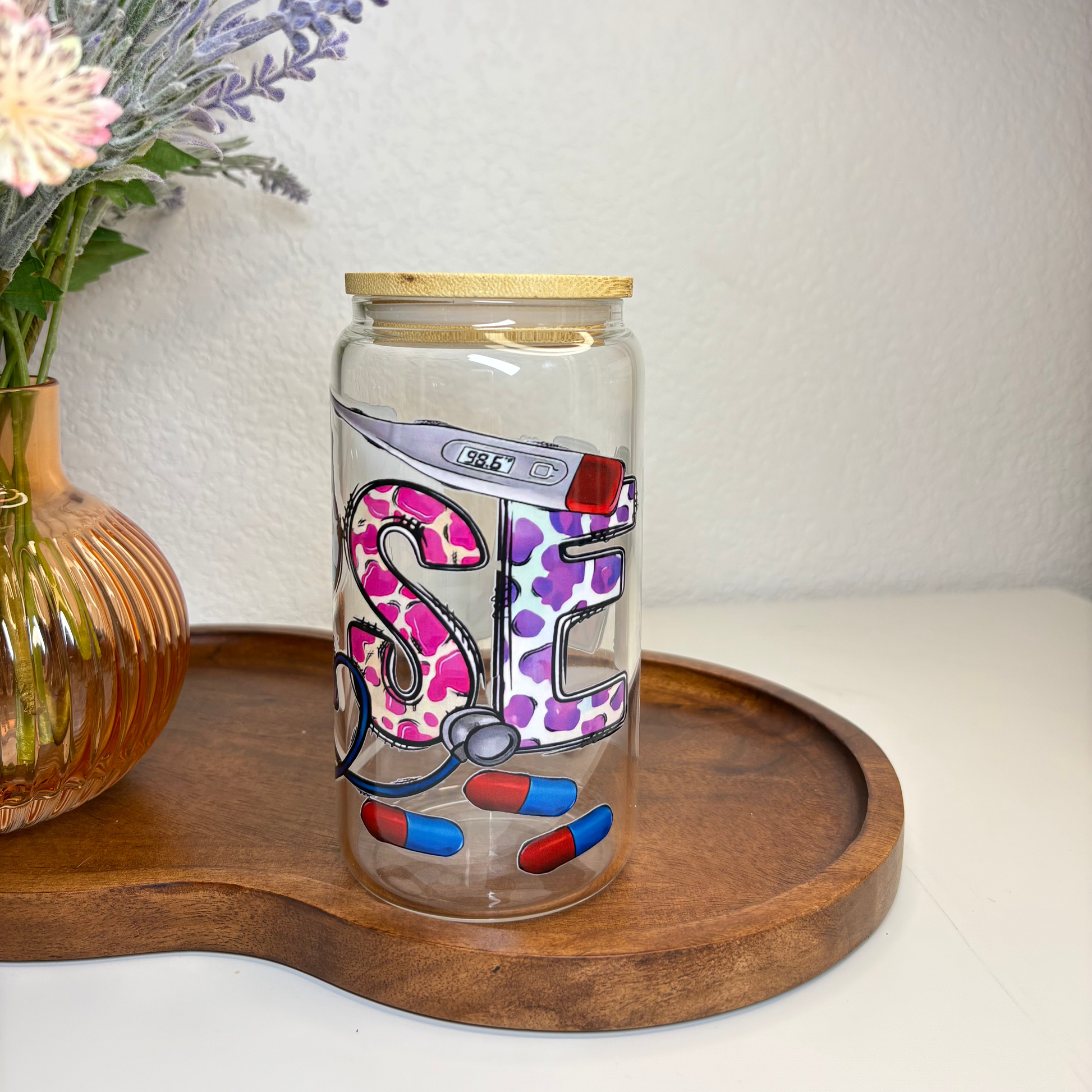 Decorative glass jar with colorful lettering on a wooden tray, next to a vase of flowers.