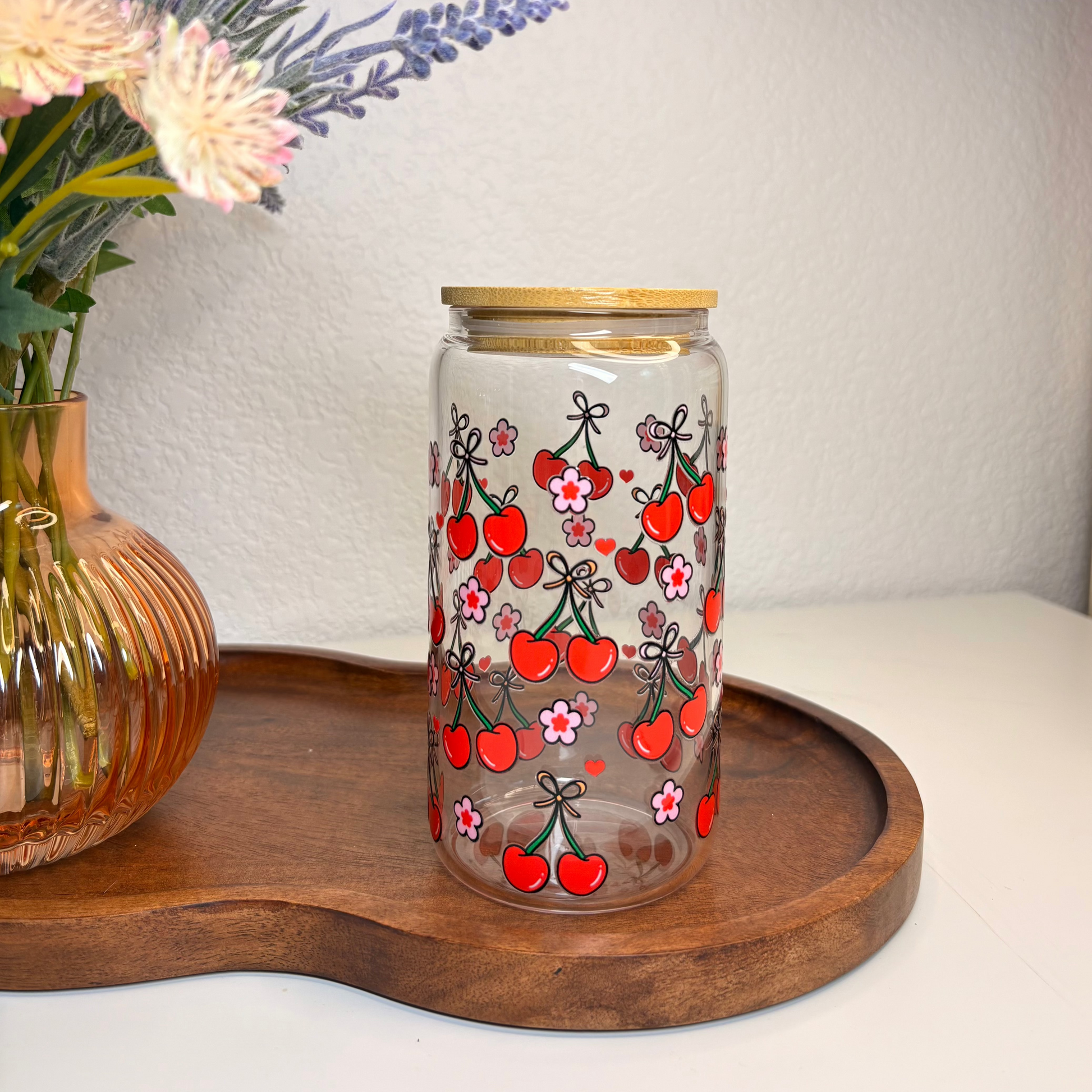Decorative glass jar with cherry design on a wooden tray with flowers in the background